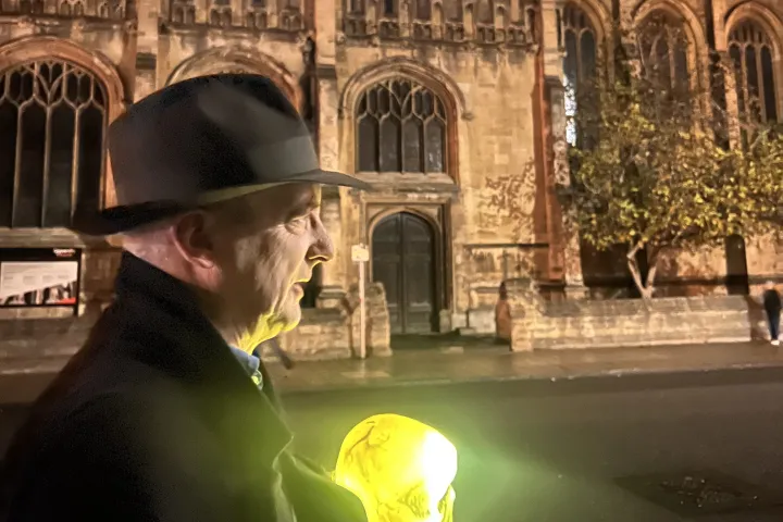 Man in hat holding glowing skull in front of illuminated gothic building at night.