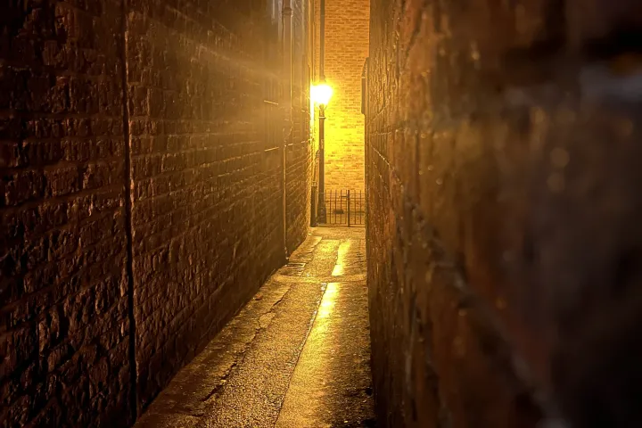 Narrow dimly lit alley with wet pavement and a glowing streetlamp at night.