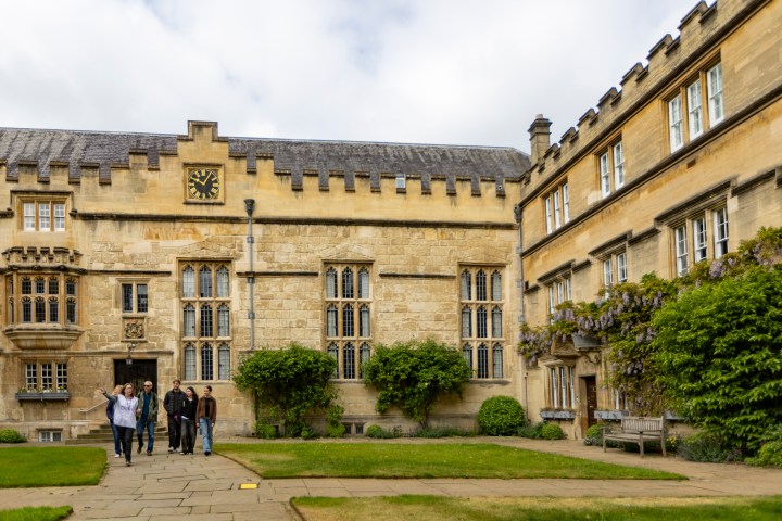 People walking in front of an old stone building with large windows and a clock.