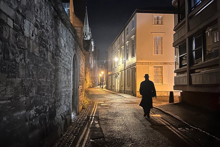 Silhouette of person in hat walking down a dimly lit cobblestone street at night.