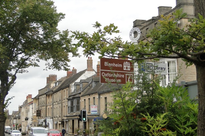 Street scene with old buildings and direction signs for Blenheim and Oxfordshire Museum amid greenery.