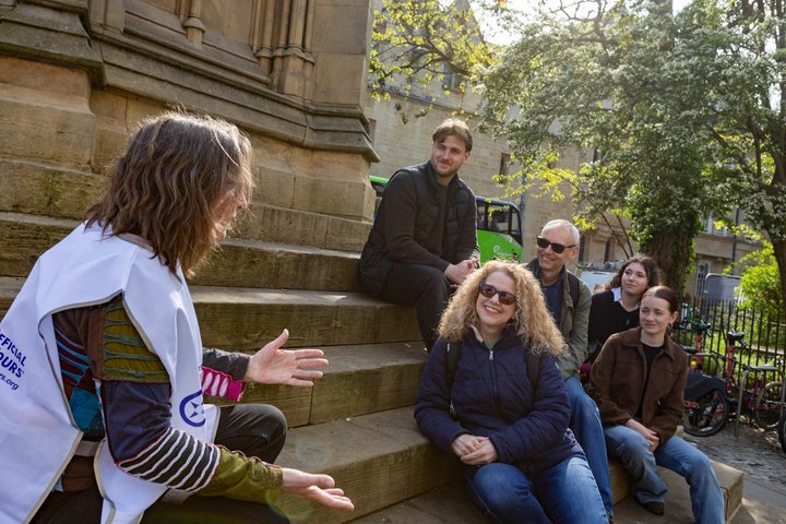 Group sits on steps, listening to a woman gesturing while speaking.