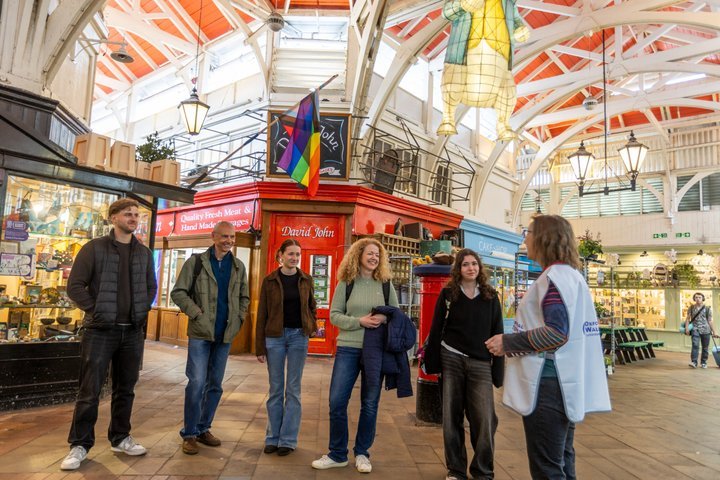 A group of people in a market with colorful stalls and a pride flag.