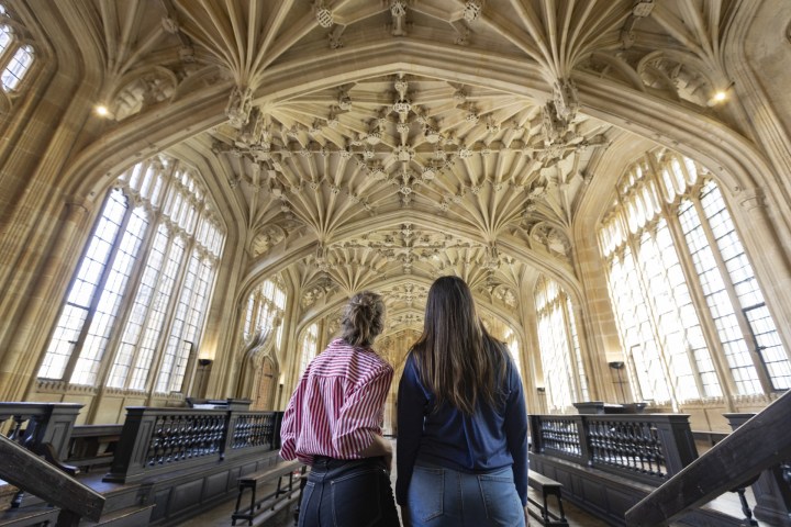 Two people in a grand hall with ornate vaulted ceiling and large windows.