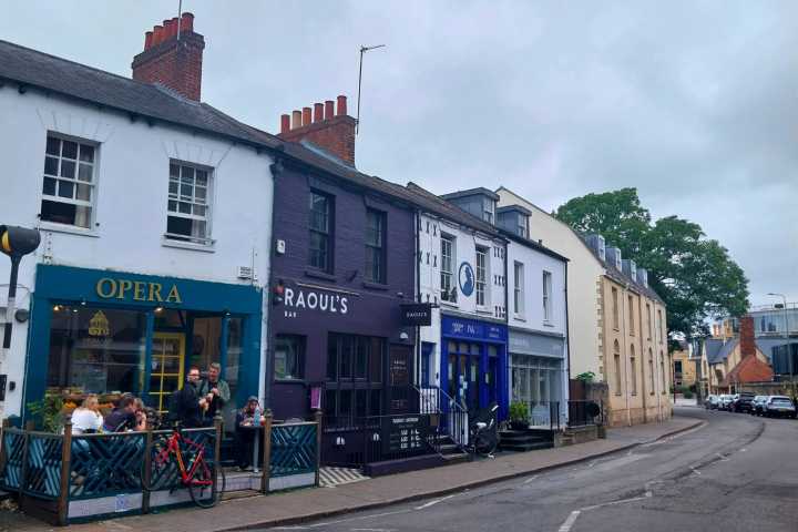 Street with row of colorful shops and cafes, people sitting outside, cloudy sky.