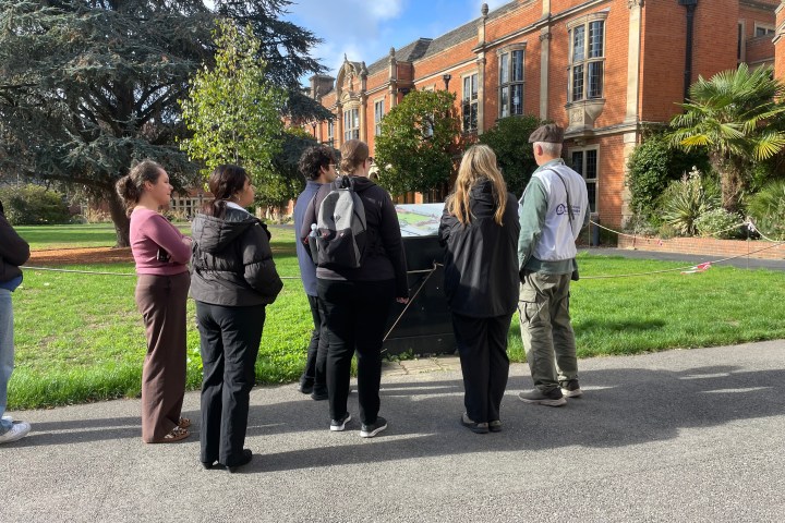 Group of people standing outside a brick building, looking at a map or information board.